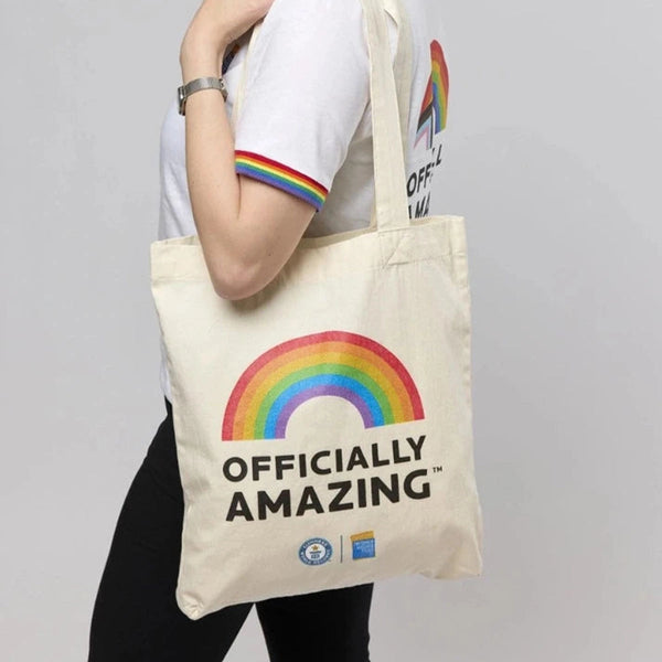 Person holding a tote bag with a rainbow and 'Officially Amazing' text on a plain background