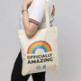 Person holding a tote bag with a rainbow and 'Officially Amazing' text on a plain background
